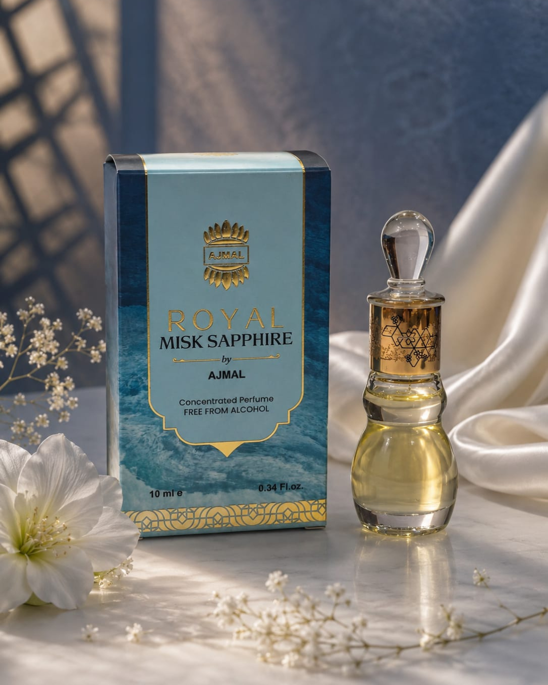 Professional luxury product photoshoot of Ajmal Royal Misk Sapphire concentrated perfume featuring a blue and gold perfume box alongside an elegant glass attar bottle. The fragrance is displayed on a marble surface with soft white satin fabric, delicate white flowers, and warm studio lighting, creating a premium and sophisticated presentation.