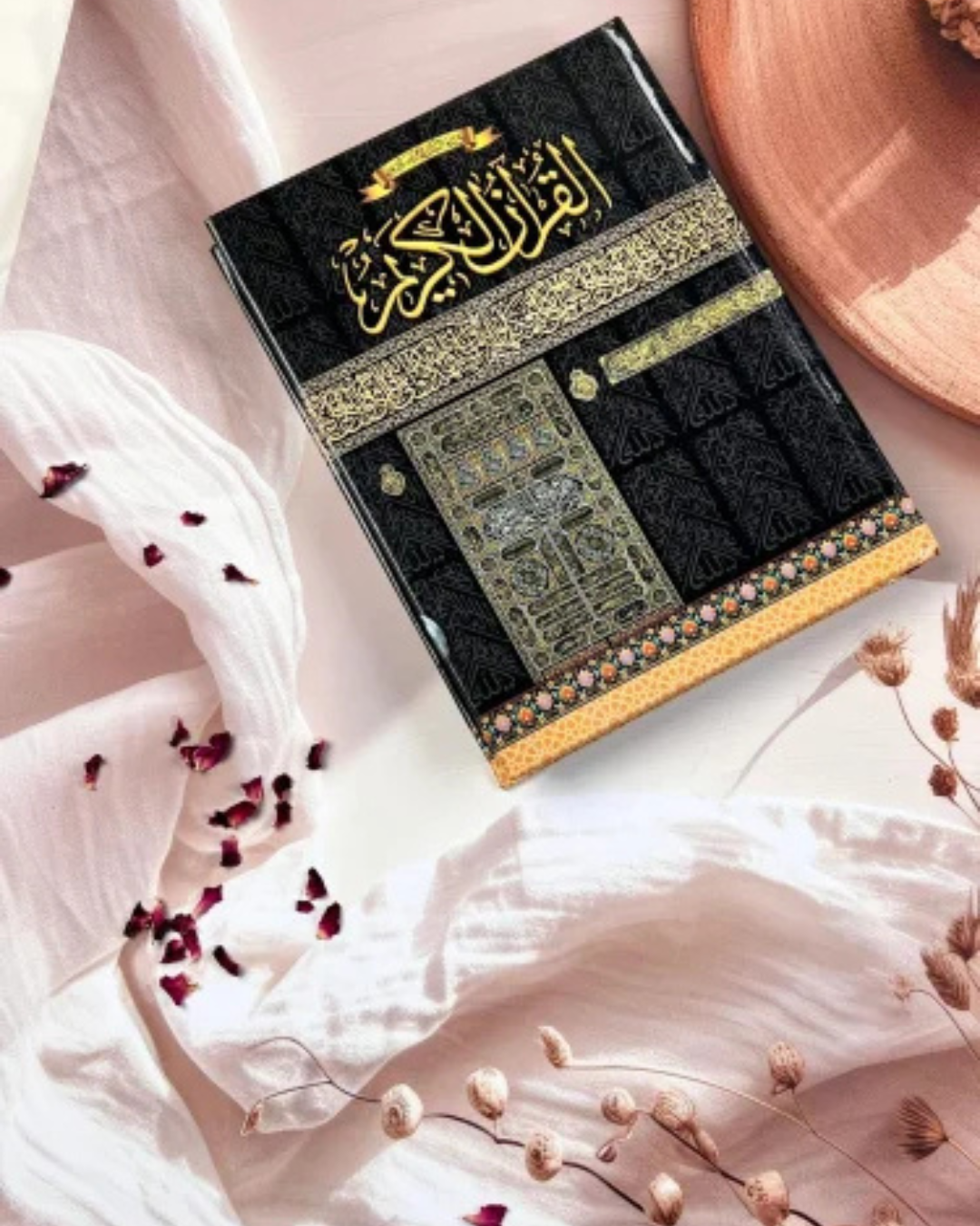 Beautifully designed Holy Quran with an ornate black and gold cover featuring Arabic calligraphy, placed on soft white fabric with scattered rose petals and dried floral accents. The elegant flat-lay arrangement creates a serene and luxurious Islamic gift presentation